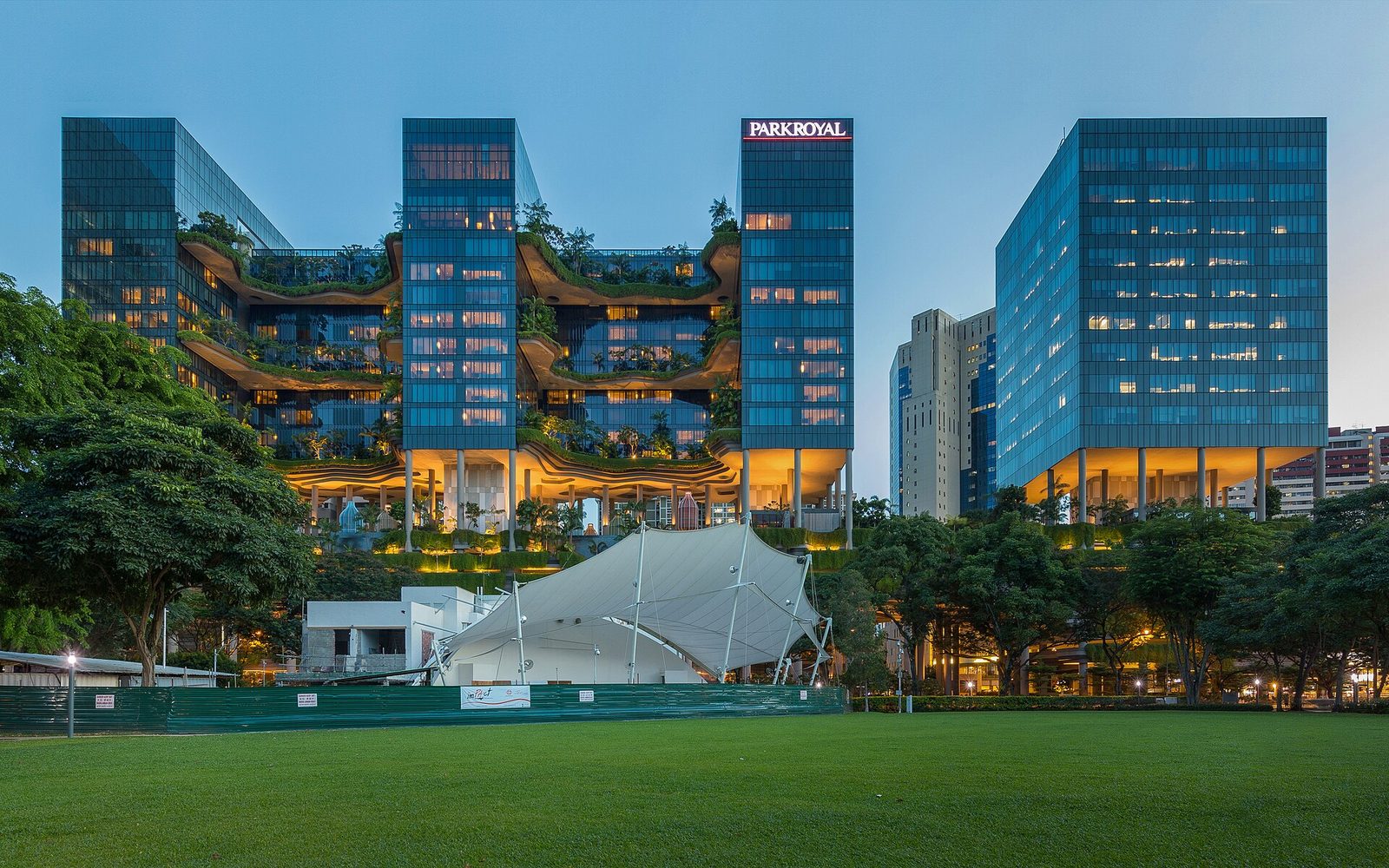 Pelouse de Hong Lim Park au crépuscule, avec un auvent blanc et la façade végétalisée de l'hôtel Parkroyal Collection Pickering illuminée en arrière-plan