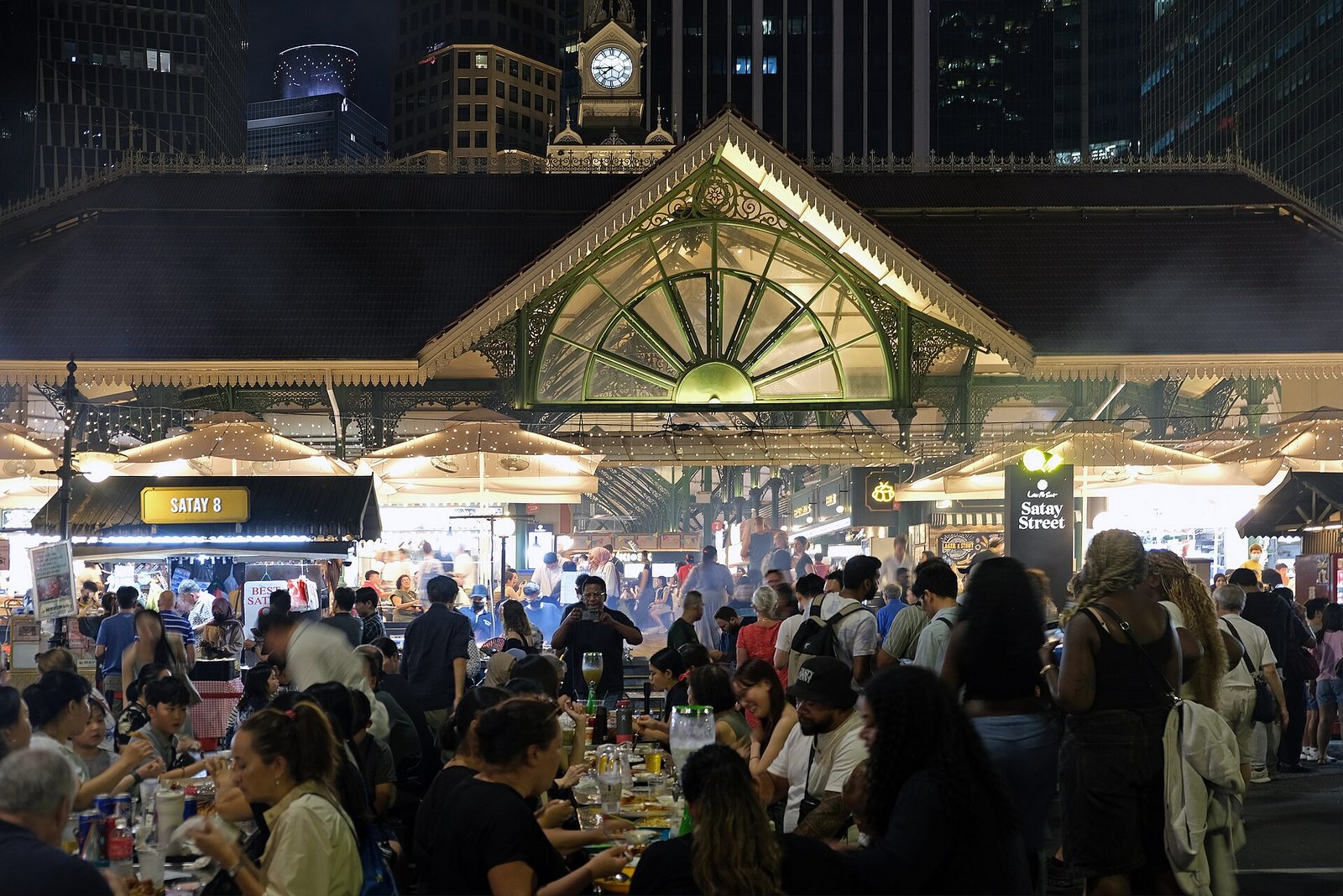 Les tablées de Satay Street, la nuit, devant la halle illuminée de Lau Pa Sat et sa tour de l'horloge, entourées de gratte-ciel