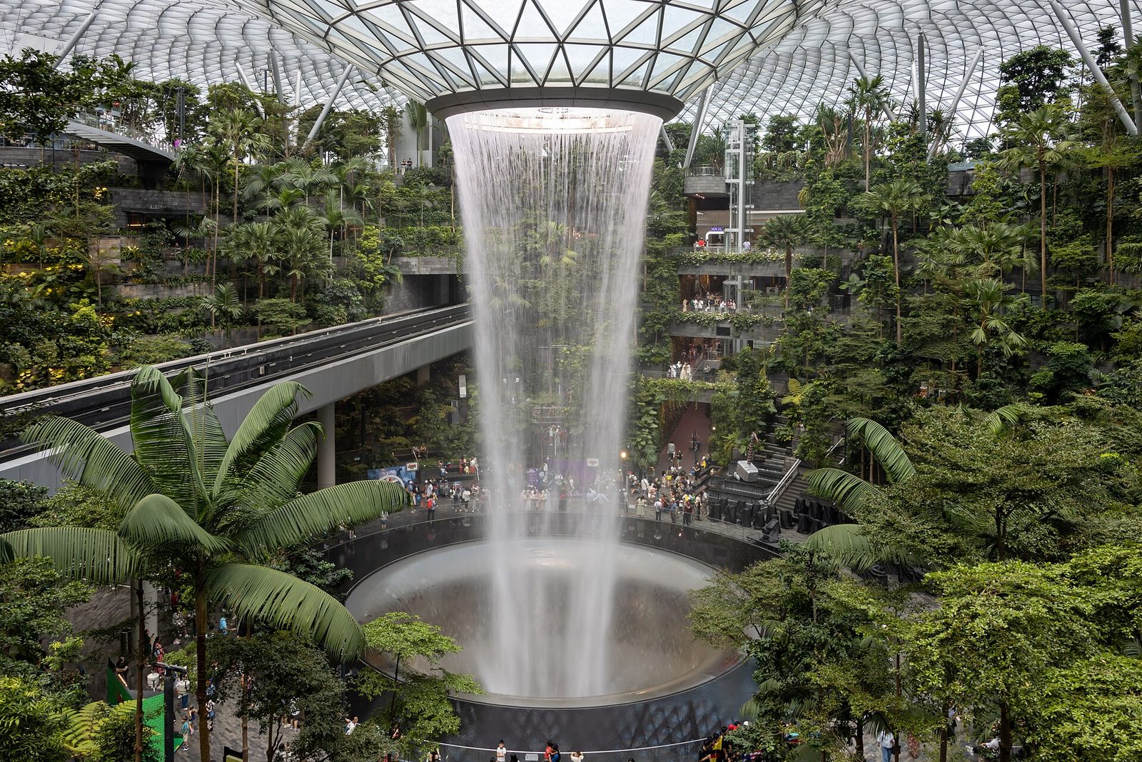 Le Rain Vortex de Jewel Changi Airport, colonne d'eau tombant d'une ouverture du dôme de verre au milieu d'une forêt intérieure, avec le Skytrain qui passe à côté