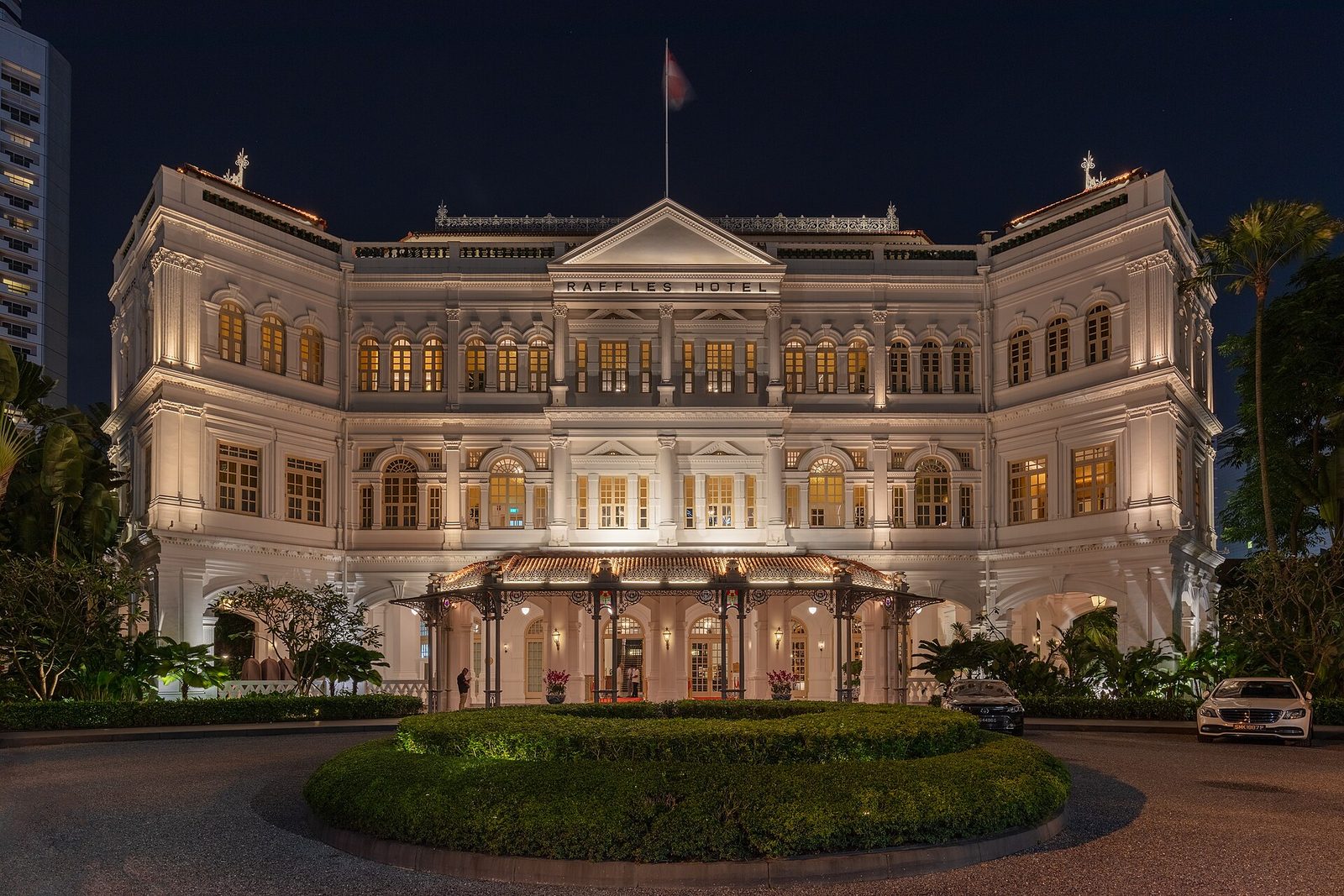 La façade blanche à colonnades de l'hôtel Raffles illuminée de nuit, avec son allée d'entrée et sa pelouse circulaire