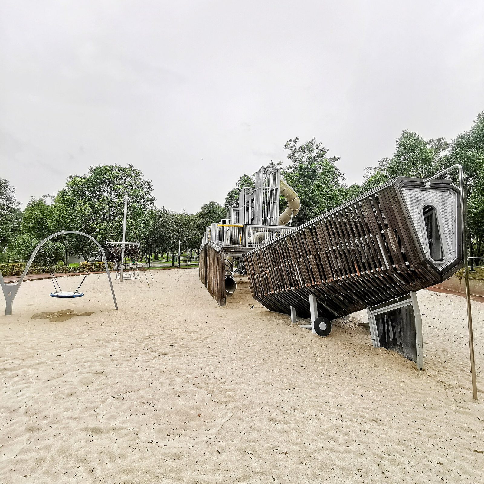 L'aire de jeux en forme de navire de guerre de Sembawang Park, coque en bois et acier posée dans un grand bac à sable, avec une balançoire nid au premier plan