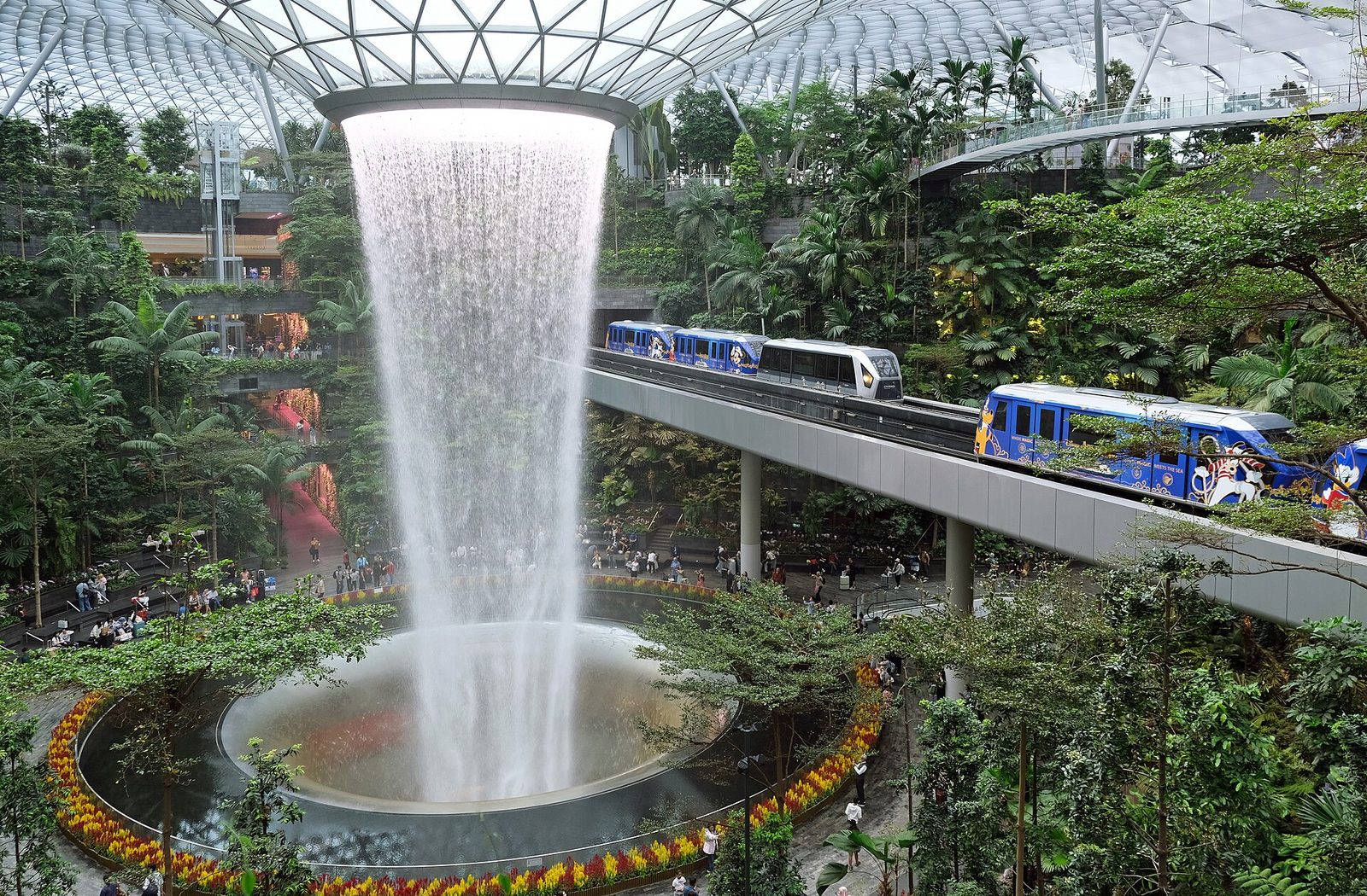 La cascade du Rain Vortex tombe du centre du dôme de verre de Jewel, entourée de végétation, avec le Skytrain de l'aéroport qui passe à droite