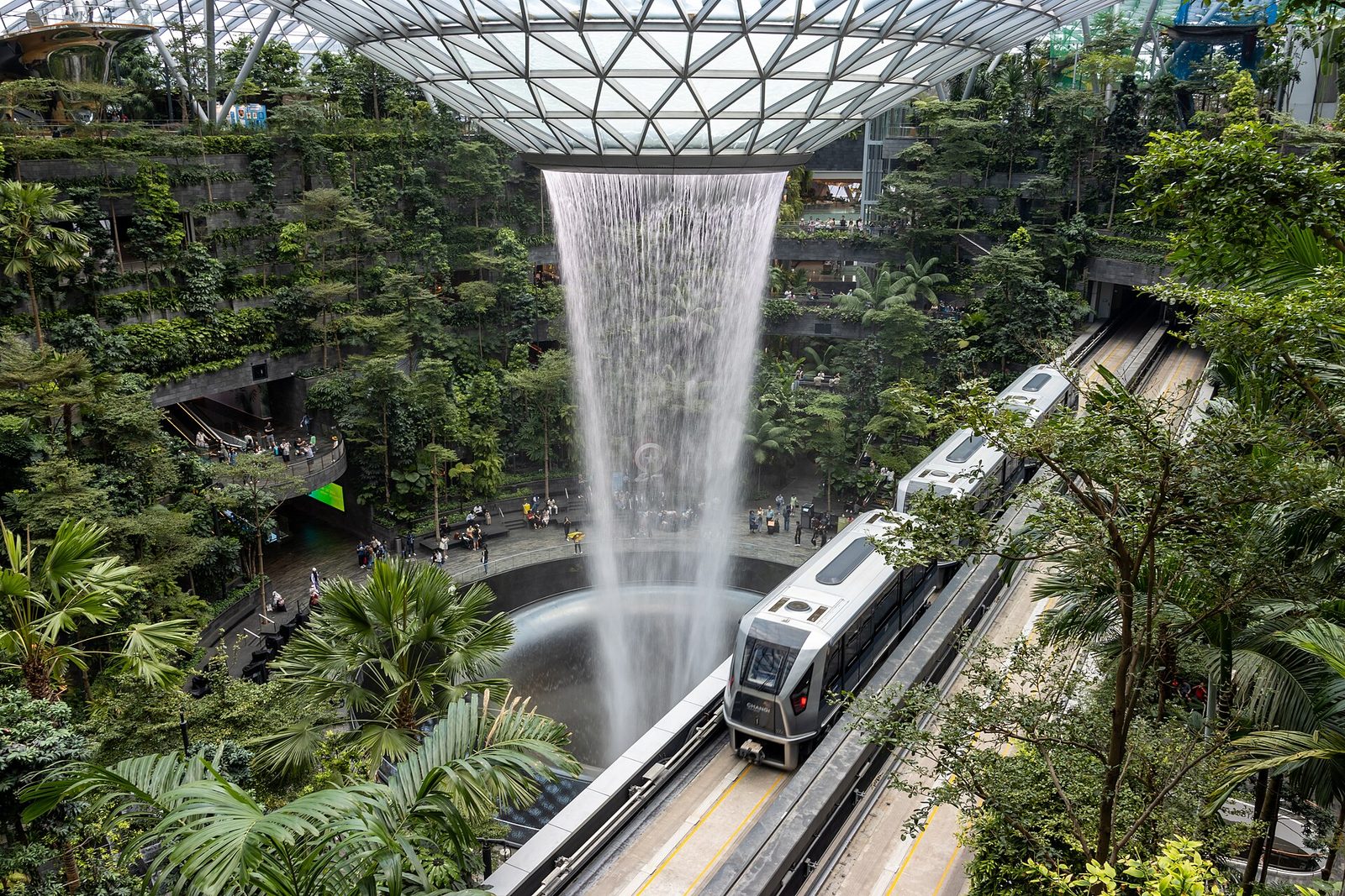 La cascade intérieure Rain Vortex de Jewel, à l'aéroport de Changi, entourée de végétation, avec une rame du Skytrain au premier plan