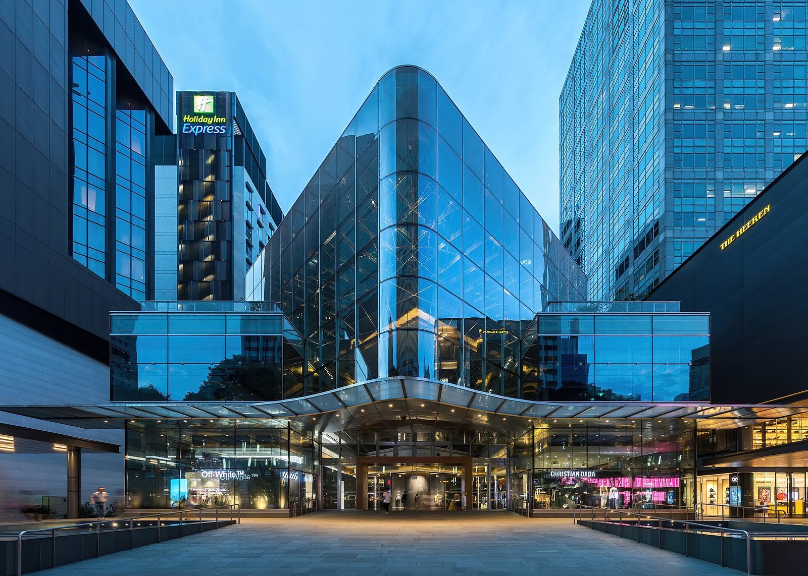 Façade de verre illuminée d'un centre commercial d'Orchard Road à l'heure bleue, avec des tours de verre de part et d'autre