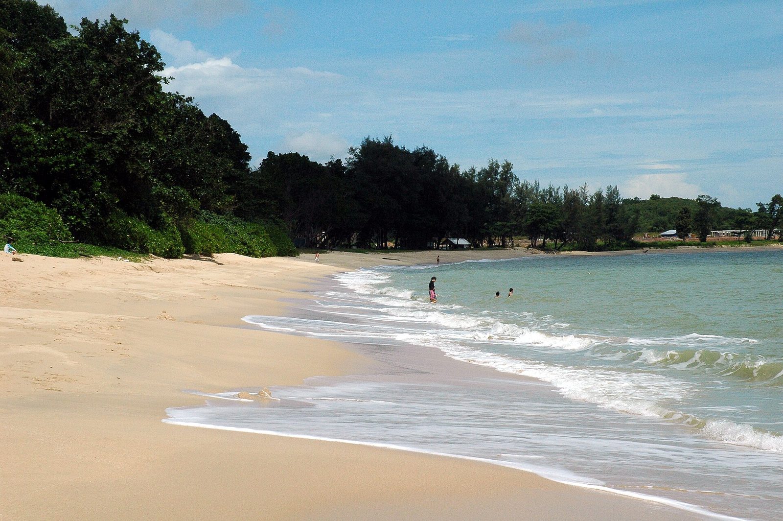 Longue plage de sable clair bordée d'arbres à Desaru, avec quelques baigneurs au loin et des vagues sous un ciel bleu