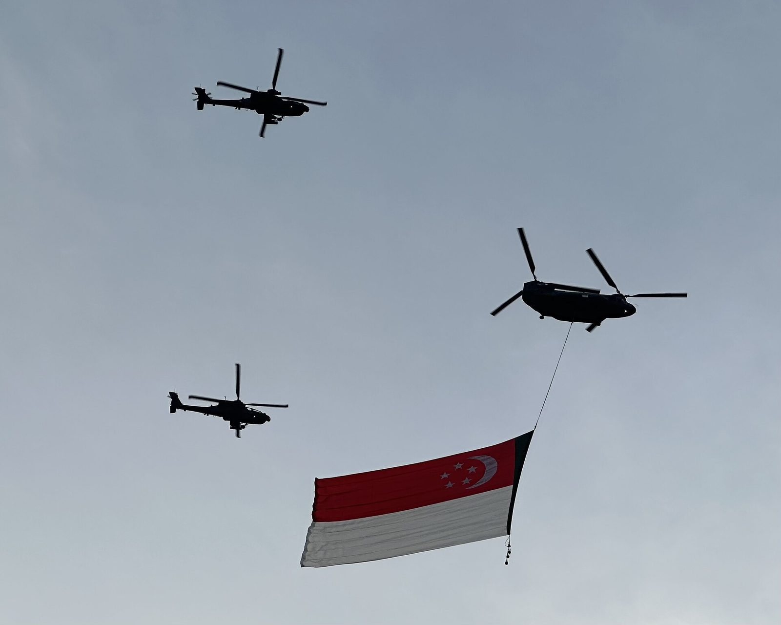 Un hélicoptère Chinook transporte un immense drapeau singapourien rouge et blanc, escorté de deux hélicoptères Apache, dans un ciel gris