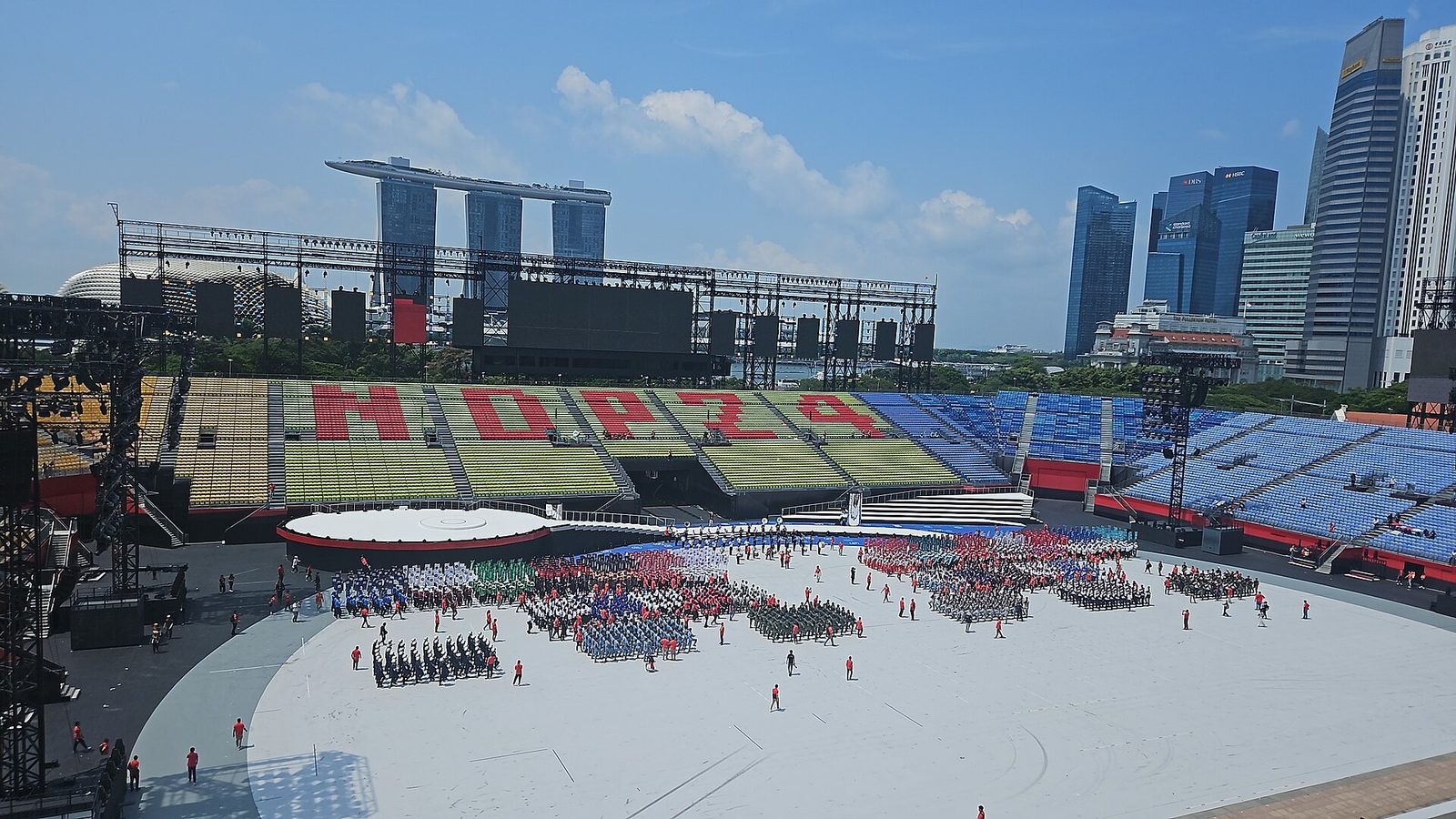 Vue en hauteur du Padang aménagé pour la parade : tribunes colorées formant les lettres NDP 24, contingents alignés sur la pelouse, tours de Marina Bay Sands au fond
