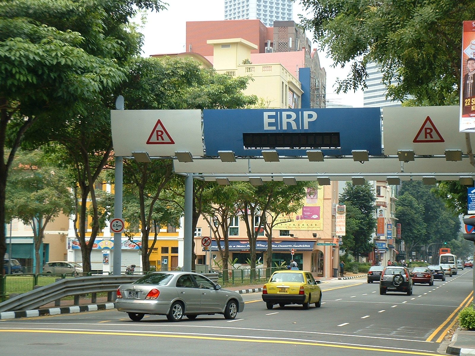 Un portique ERP bleu et blanc au-dessus d'une avenue bordée d'arbres et de shophouses colorées, avec un taxi jaune et une voiture grise