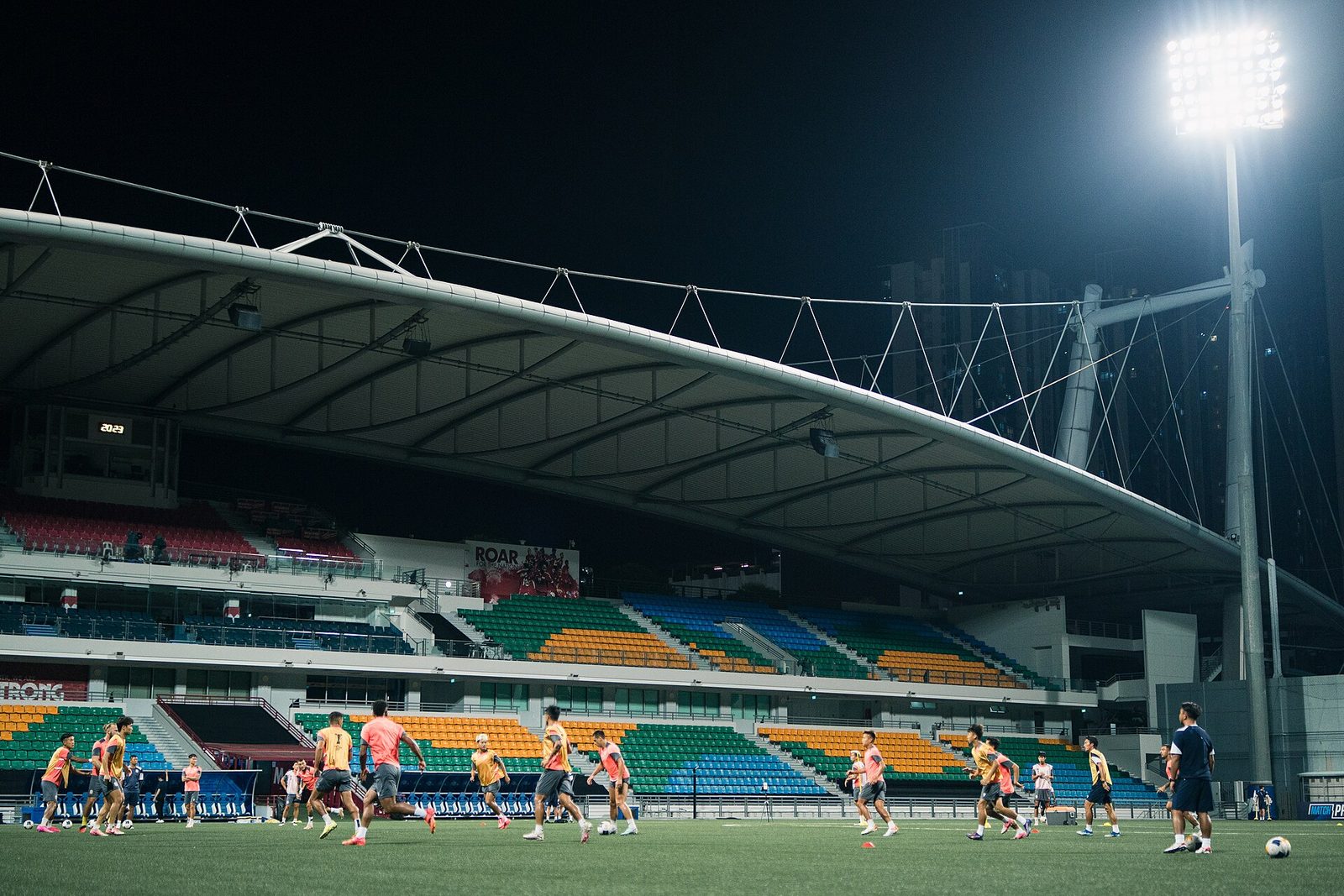 Le stade de Jalan Besar éclairé de nuit, avec sa tribune couverte aux sièges colorés et des joueurs sur la pelouse