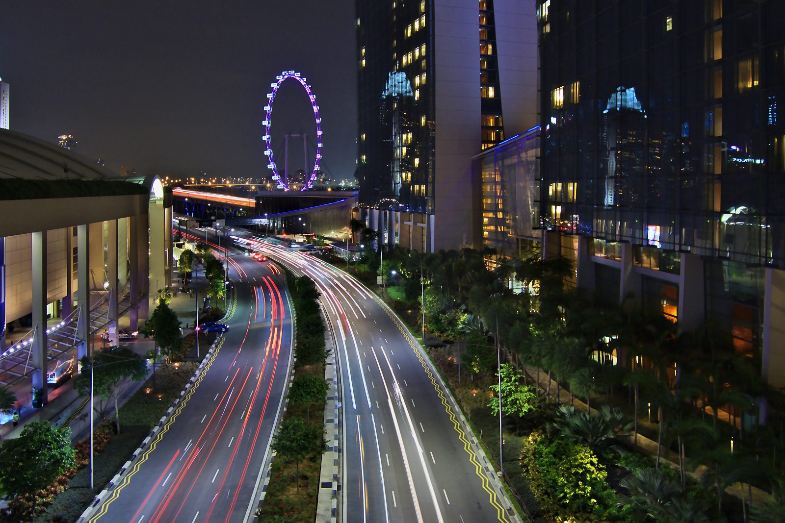 Traînées lumineuses des voitures sur une route courbe de nuit, entre l'hôtel Marina Bay Sands et la grande roue Singapore Flyer éclairée en violet