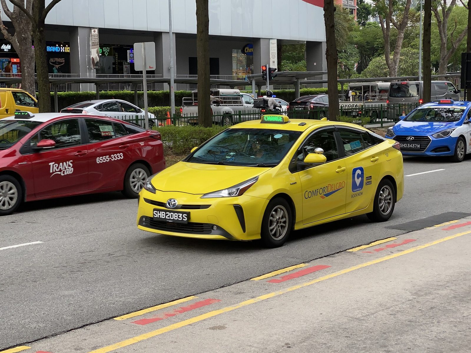 Un taxi ComfortDelGro jaune roule entre un taxi Trans-cab rouge et un taxi bleu, sur une avenue bordée d'arbres