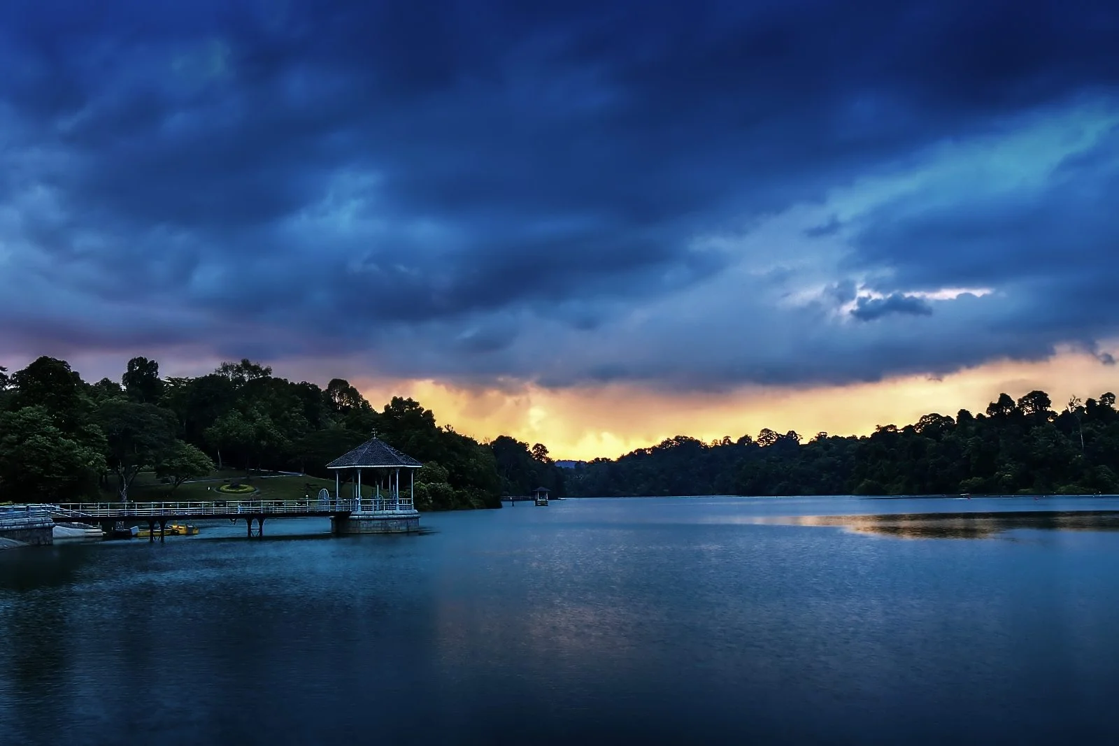 Ciel d'orage bleu sombre au coucher du soleil au-dessus du réservoir MacRitchie, avec un kiosque au bord de l'eau et la forêt sur les rives