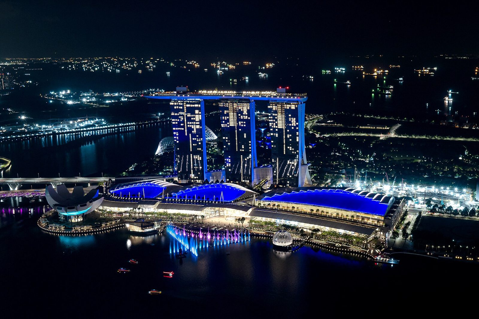 Vue aérienne de nuit de Marina Bay Sands illuminé en bleu, avec les jets d'eau colorés du spectacle Spectra au bord de la baie