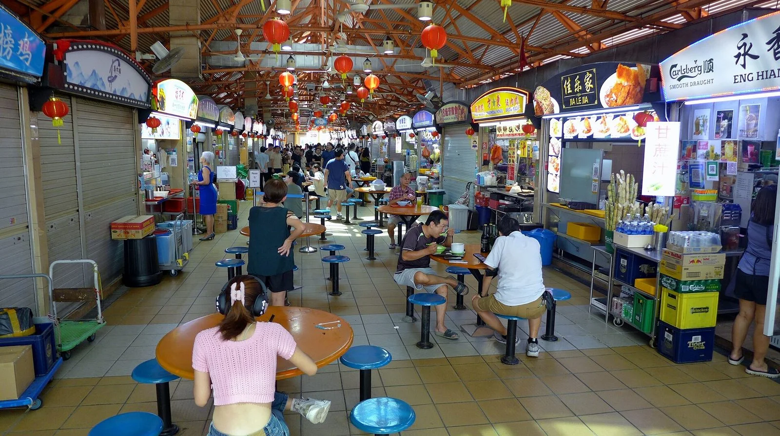 Allée centrale du Maxwell Food Centre bordée de stands, avec des tables rondes et des tabourets bleus, sous des lanternes rouges