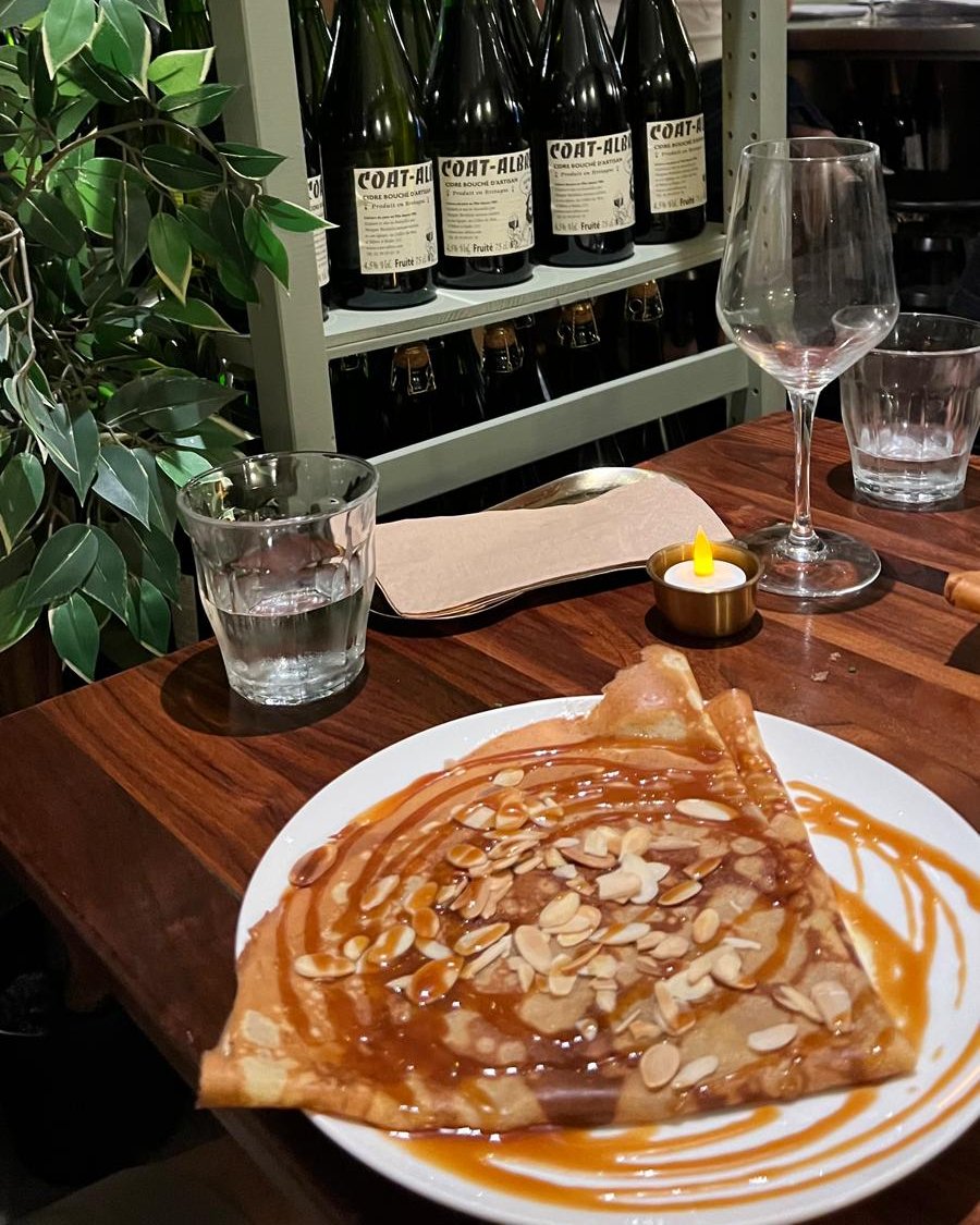Crêpe pliée nappée de caramel et d'amandes effilées, sur une table en noyer, devant une étagère de bouteilles de cidre