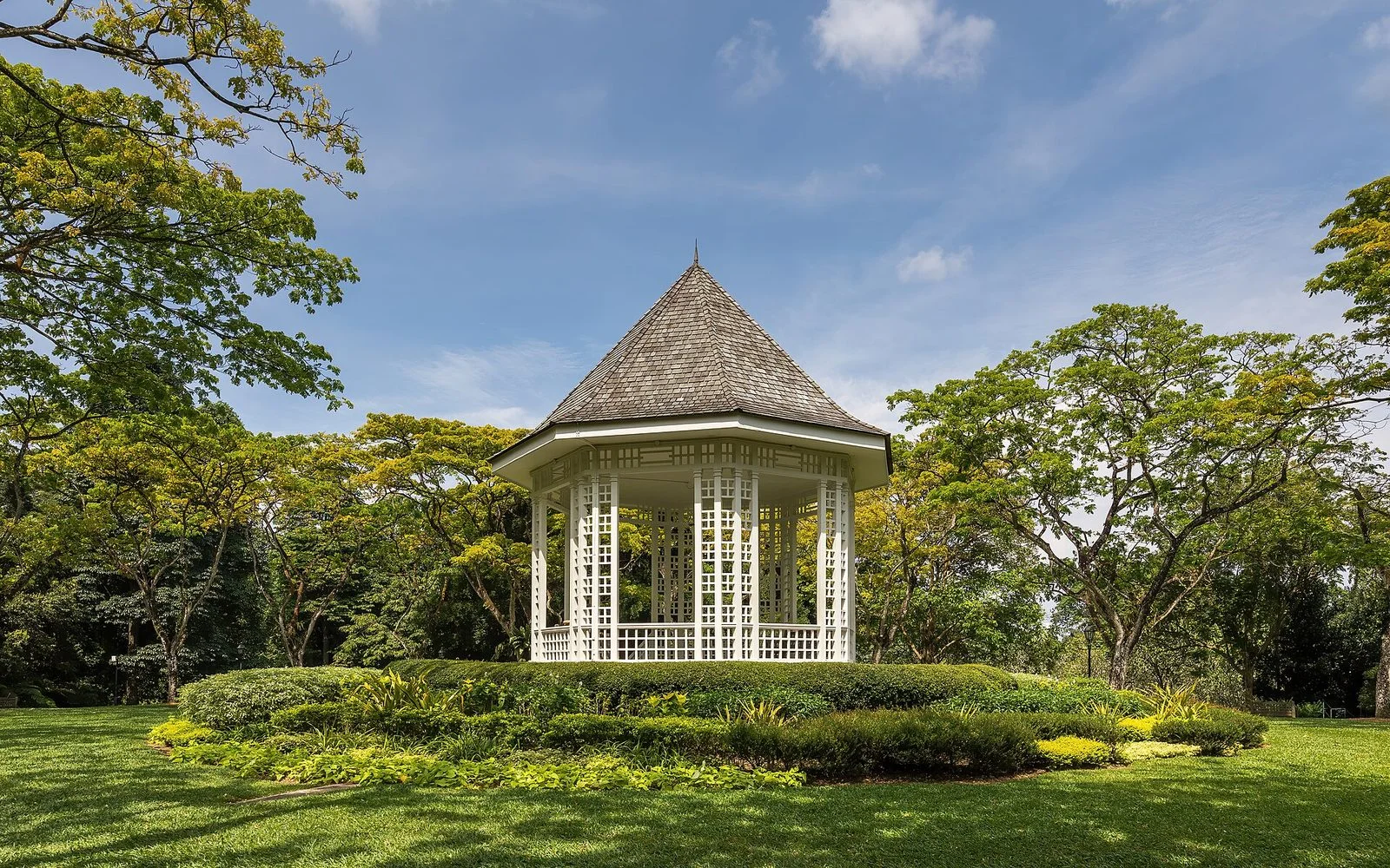 Le kiosque à musique blanc à toit pointu du jardin botanique de Singapour, entouré de pelouses et de grands arbres sous un ciel bleu